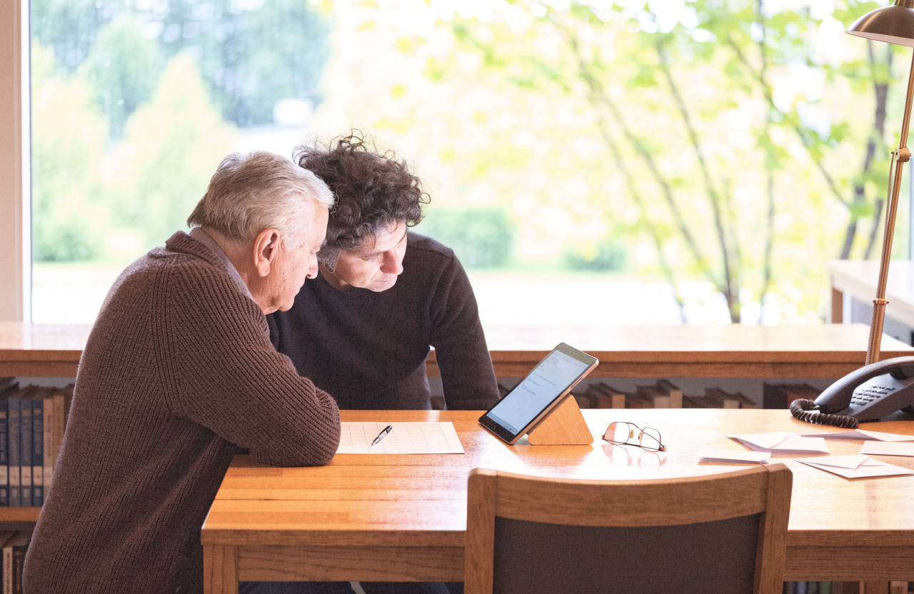An older man and a volunteer sit side by side at a library help desk, leaning over a tablet with a paper form and reading glasses beside it. The desk is a permanent fixture with its own lamp, a spare chair and a telephone to the town hall.