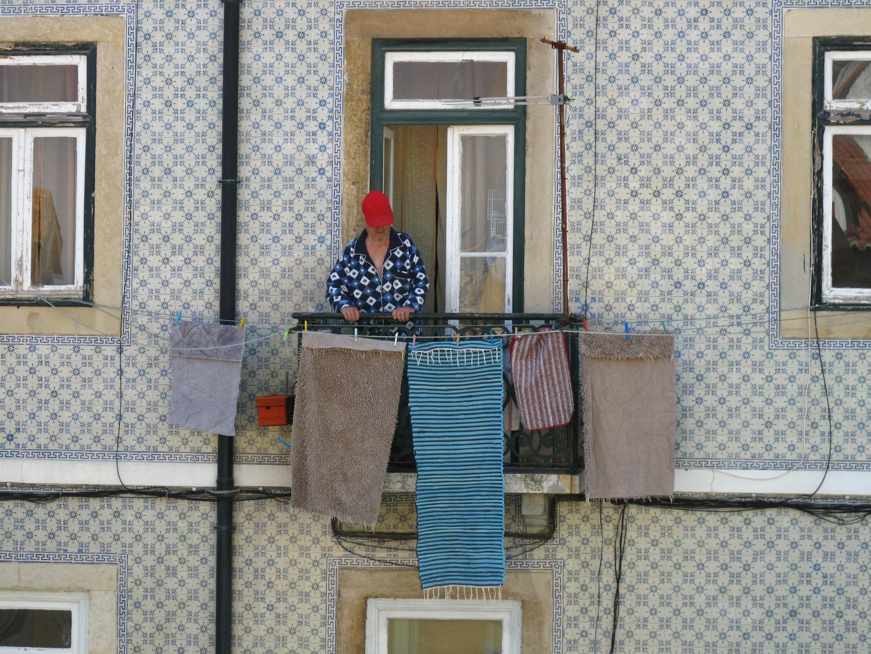 A woman stands on a Lisbon balcony with traditional Portuguese tiles, hanging laundry on a sunny day.